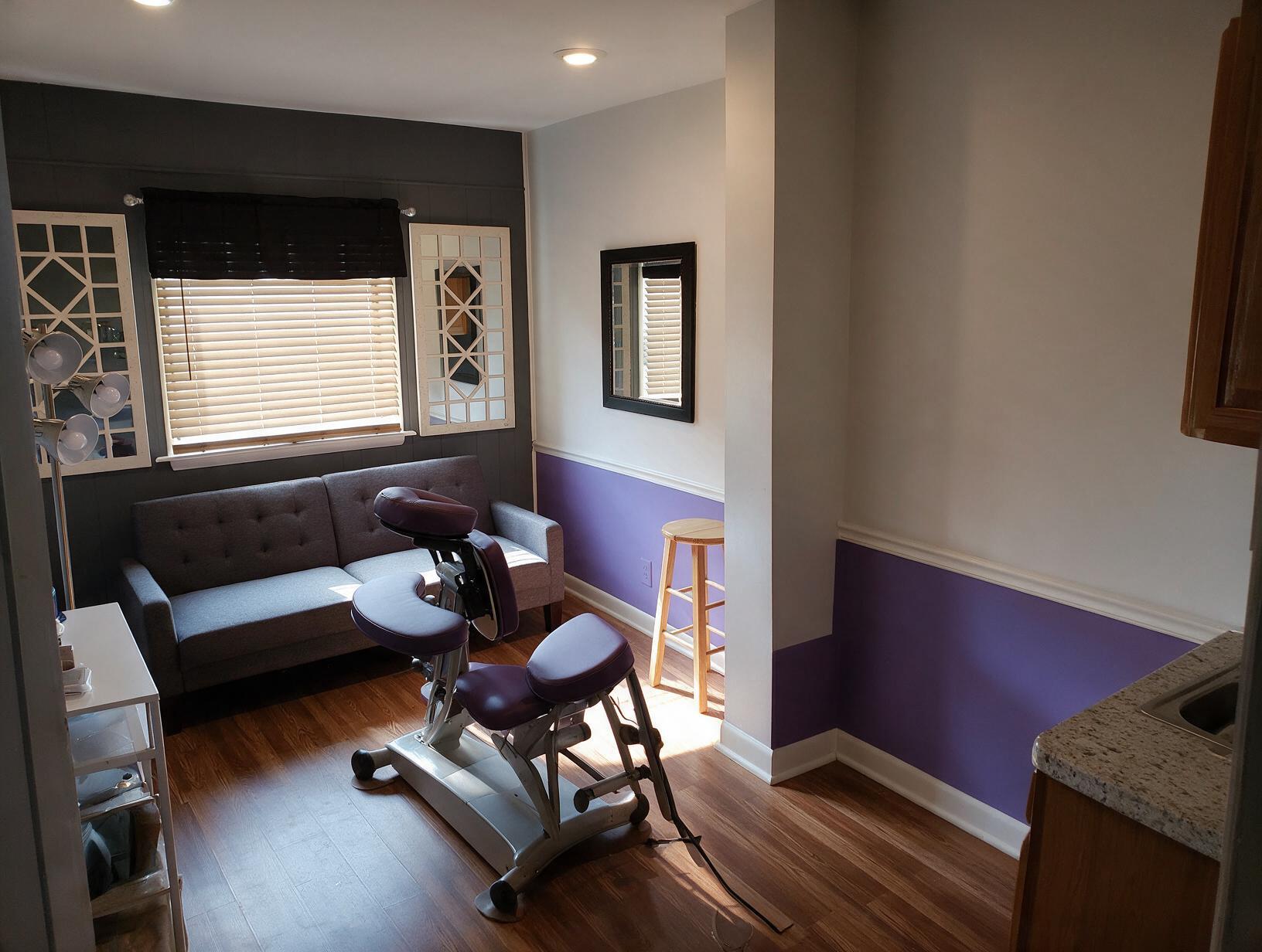 Treatment room with a purple-and-white wall, gray tufted sofa, purple massage chair, wooden stool, window blinds and mirrors.