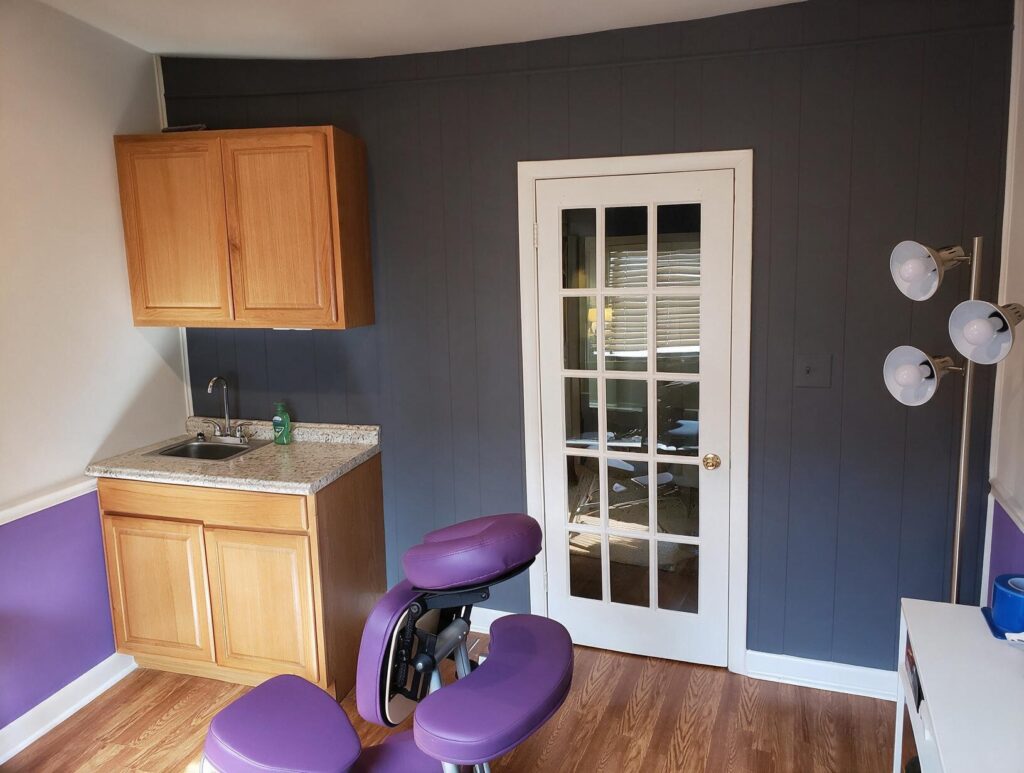 Empty treatment room with purple massage-style chair, wall cabinets and small sink, gray paneled wall and white glass door.