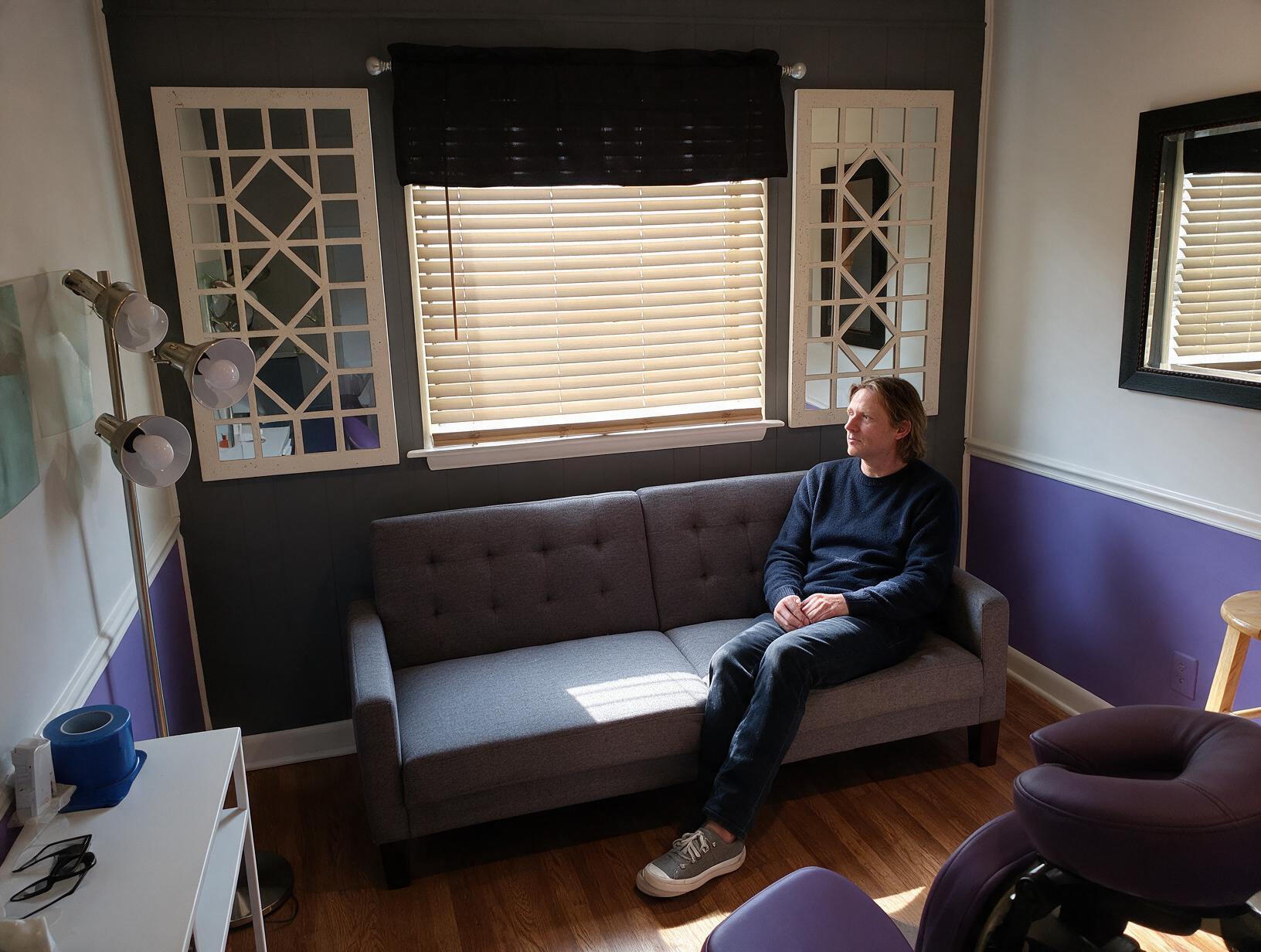 1305 inline 1791325869: carolina scalp center A man in a navy sweater and jeans sits on a grey sofa, looking out to the side, in a room with purple walls