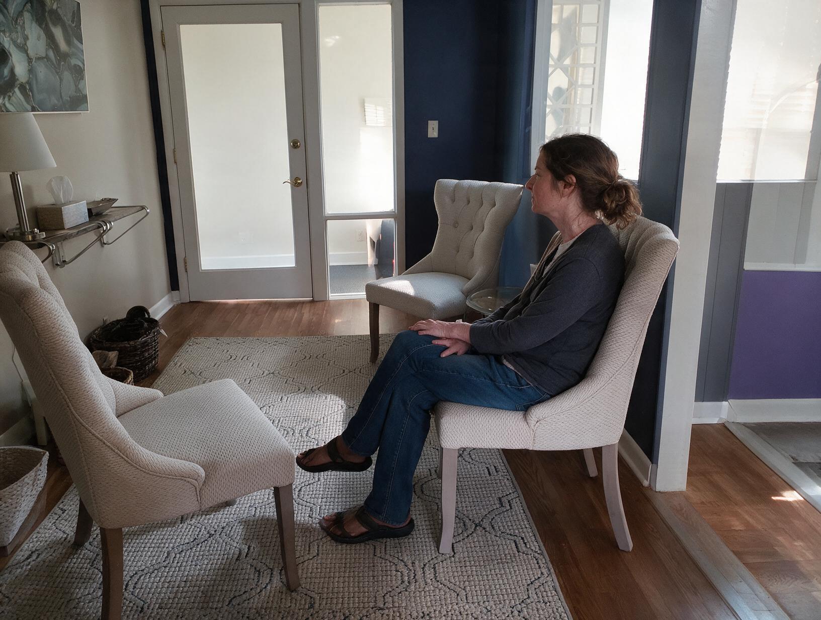 Woman in a gray cardigan and jeans sits in a cream tufted chair in a blue waiting room, looking out a window to the side.