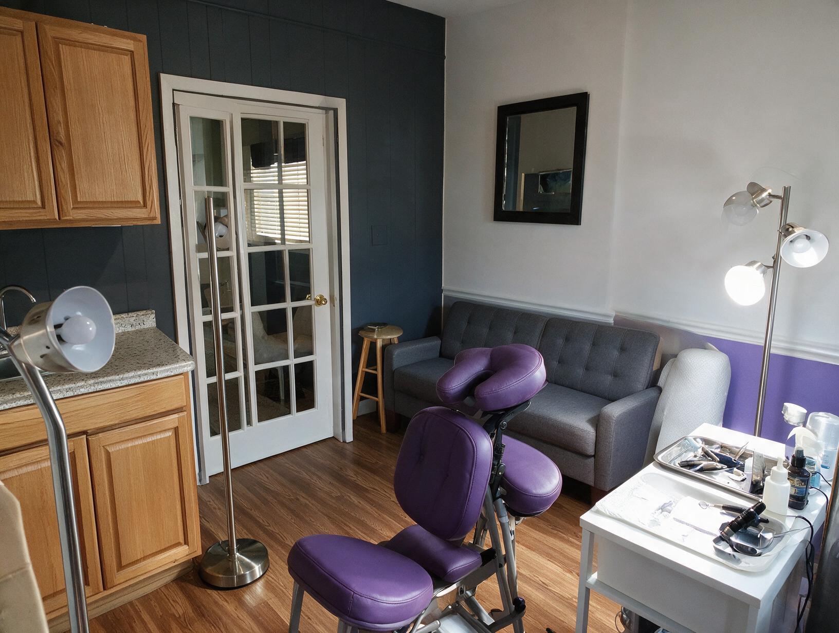 459 inline 1790980487: carolina scalp center Treatment room with a purple massage chair, grey sofa, wooden stool, floor lamps and a white tray table holding instruments.