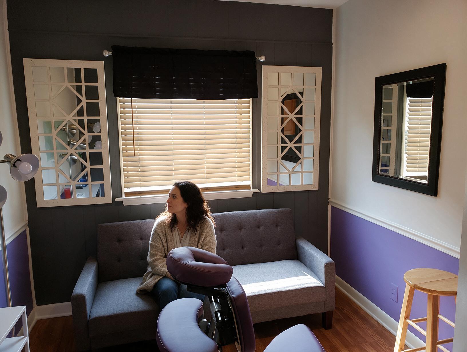Woman in a beige cardigan sits on a gray sofa, looking toward a blinded window in a room with purple and white walls.
