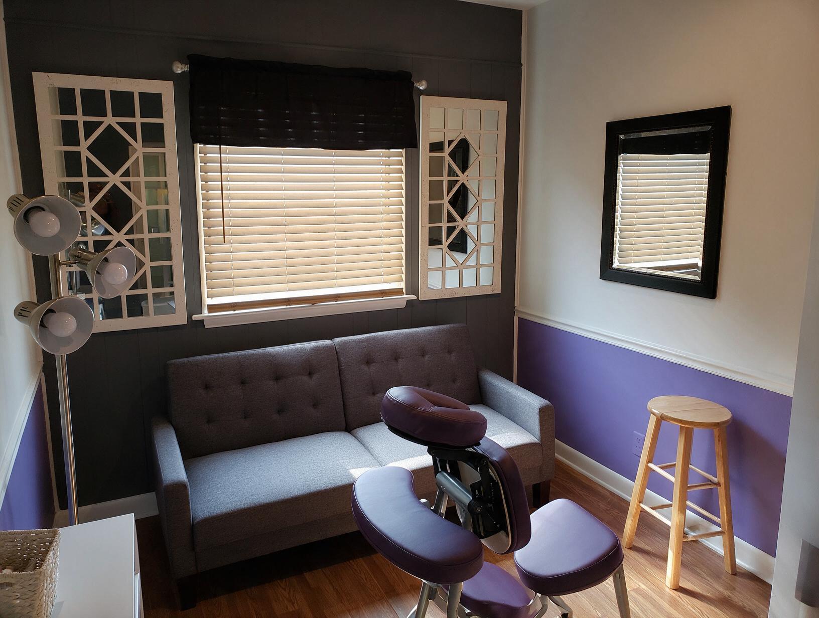 Small room with gray tufted loveseat, purple massage chair, wooden stool, mirrors, blinds and purple-and-white walls.