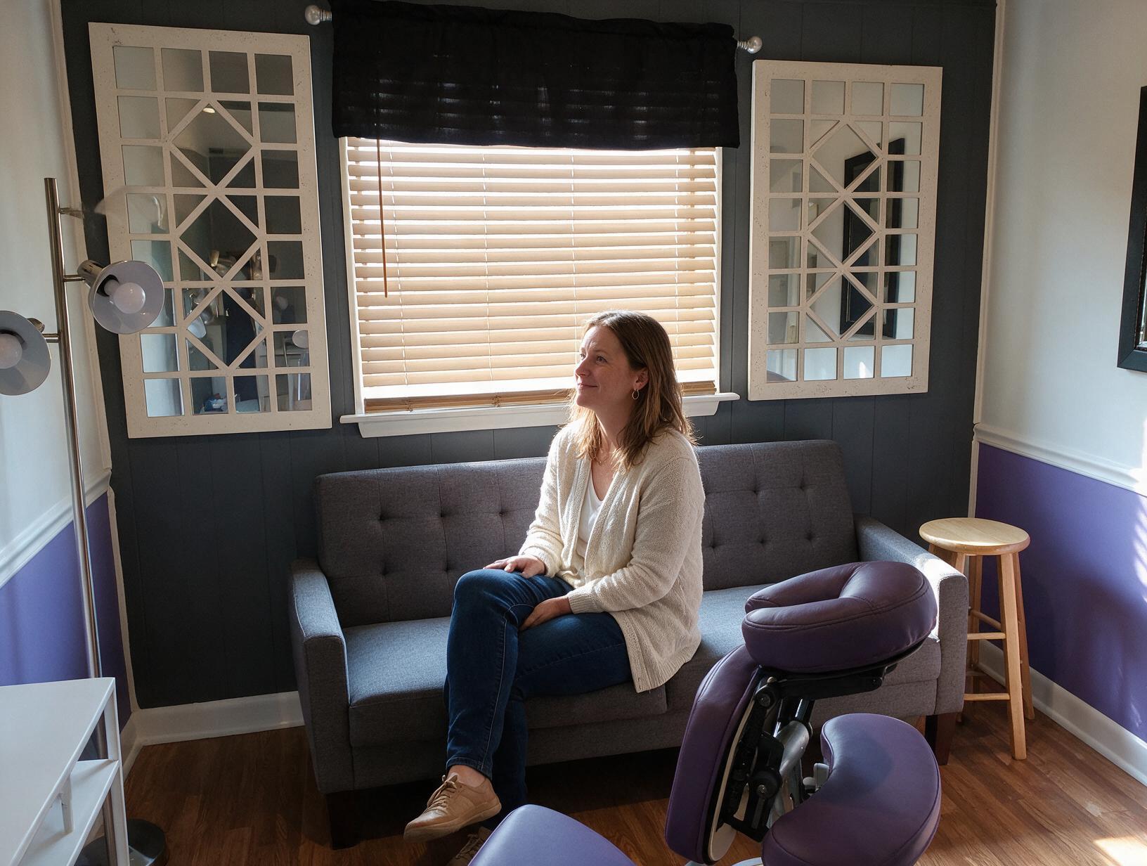 995 inline 1791066225: carolina scalp center Woman in a cream cardigan and jeans sits on a grey sofa in a purple and grey room, gazing toward a sunlit window.