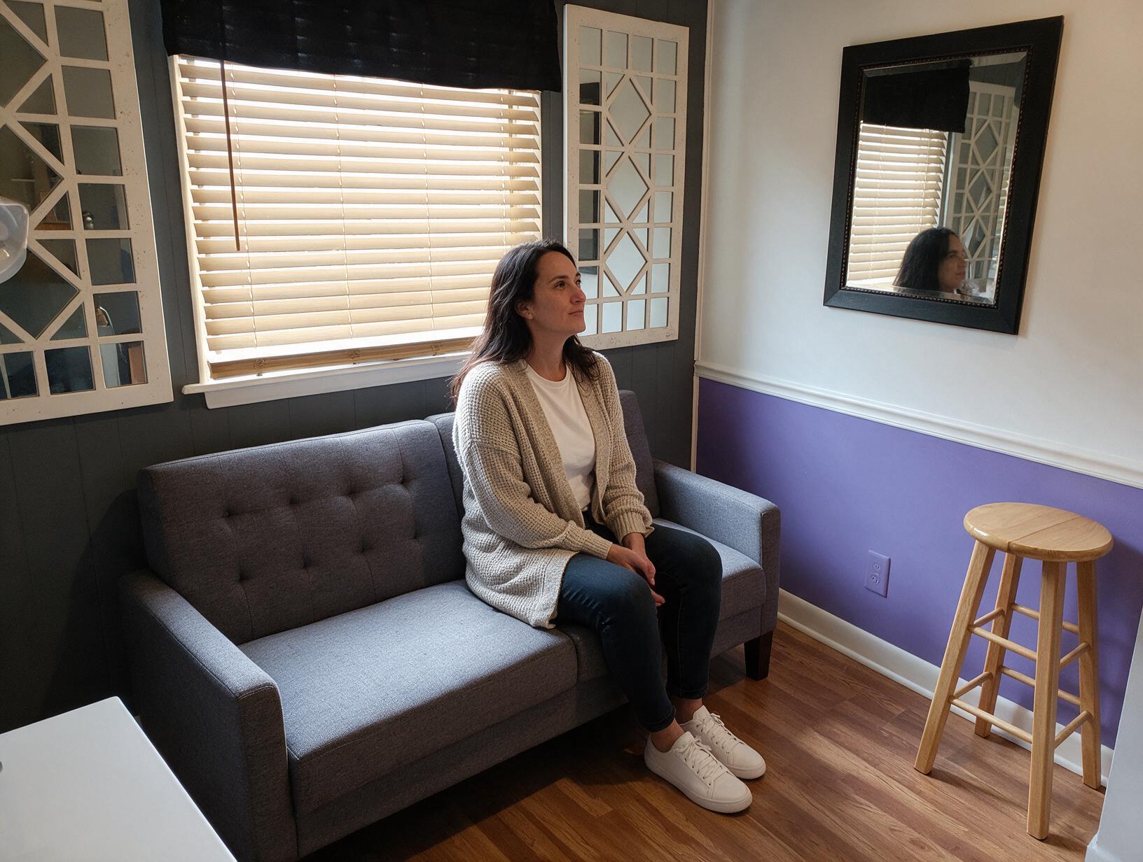 996 inline 1791035525: carolina scalp center Woman in a cream cardigan and jeans sits on a gray tufted sofa, looking toward a blinds-covered window