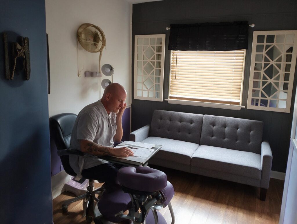 Bald tattooed man in a light grey short-sleeved shirt sits at a desk reading papers in a room with a grey sofa and sunlit