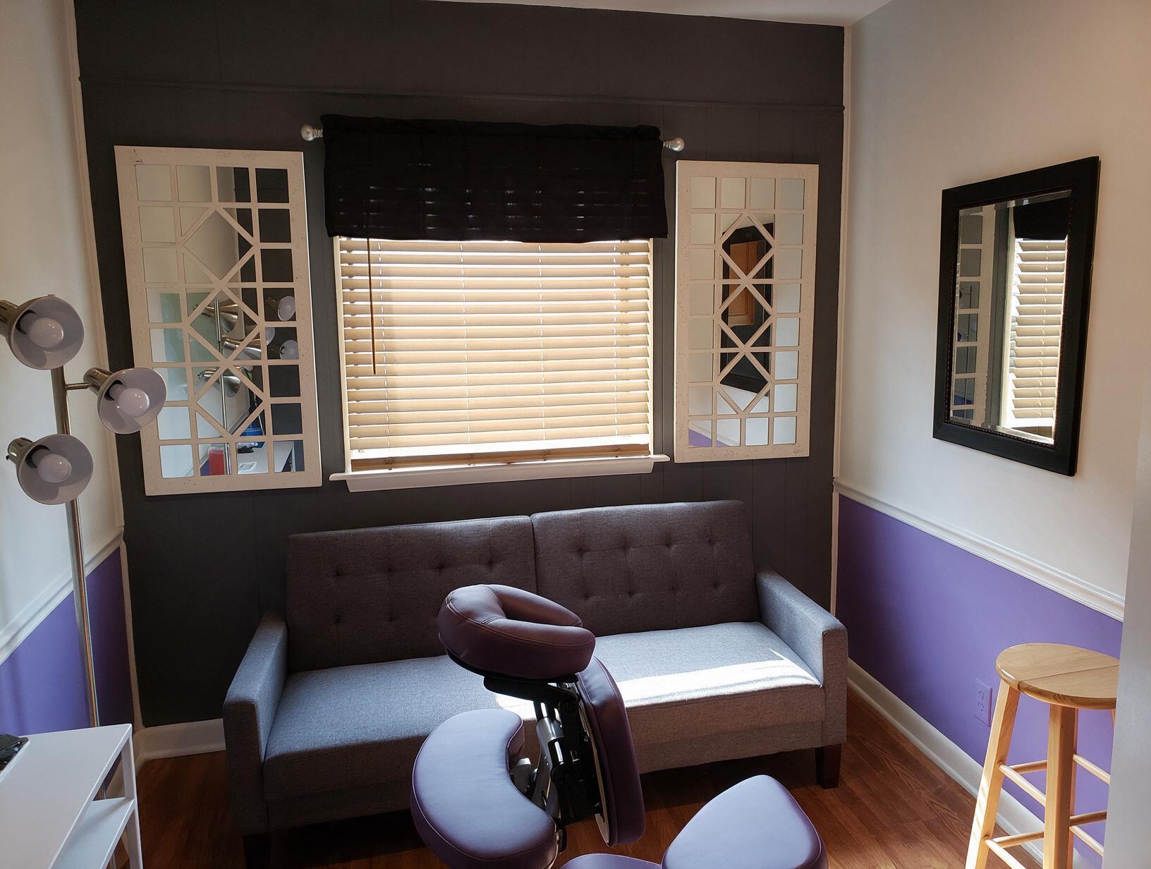 Empty treatment room with a gray sofa, purple massage chair, mirrors and blinds on a dark wall, purple and white walls